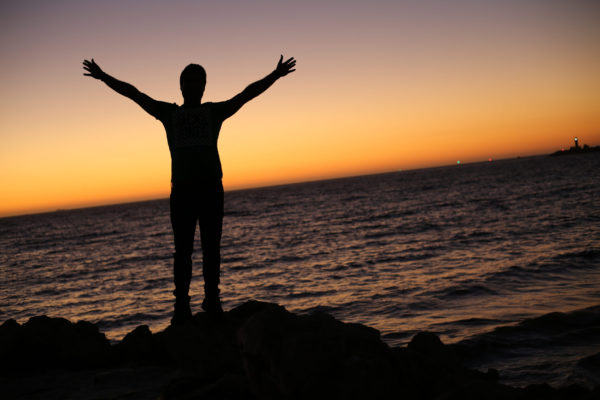 A silhouette of a person, arms raised, standing against a sunset over a lake.