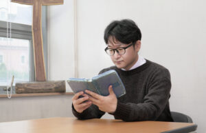 A young minister with black hair and glasses is seen seated at a table, holding and reading a bible.