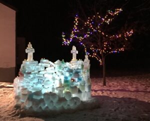 A small structure made of blue ice blocks, lit from within at night. It has two spires on the walls and a tree with Christmas lights on it behind.