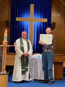 Two people standing in front of a cross and a rainbow candle. One is holding a certificate up. Both are smiling.
