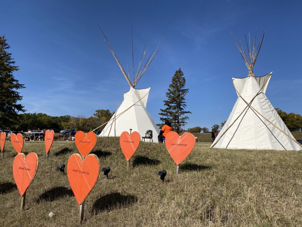 Tipis and orange hearts with names near the site of the Brandon Indian Residential School. It's late summer, and the grass is brown and the sky bright blue against the white tipis and orange hearts. 