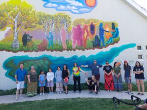 13 people posing in front of mural. These are the artists and 11 of the people portrayed in the photo.