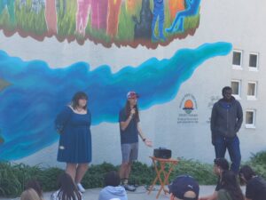 3 young people in front of the mural, one holding a microphone