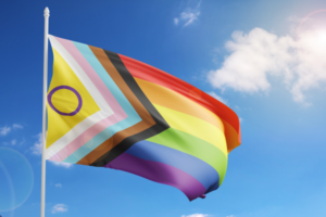 An inclusive Pride flag, with colours for trans, BIPOC, and intersex people, is flying against a blue sky.