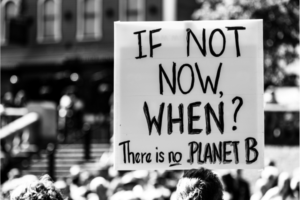 Black and white photo of a sign at a public demonstration. , hand written and held aloft by one hand. It reads, "If not now, when? There in no Planet B."