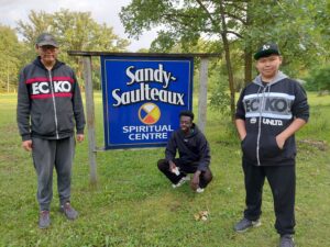 3 people in front of Sandy Saulteaux Spiritual Centre sign