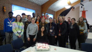 A group of people behind one person with a birthday cake