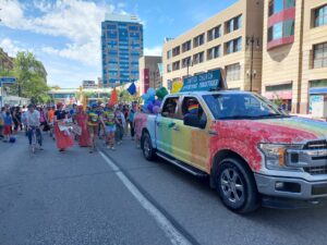 Truck decorated with rainbow colours of paint and sign saying United Church Affirming Ministries, followed by a lot of people walking