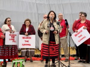 A photo of Grand Chief Cathy Merrick speaking on a small stage in front of a longhouse, on a windy and overcast day. People behind her, including the Moderator, Rev Carmen Lansdowne, hold signs reading "search the landfill".